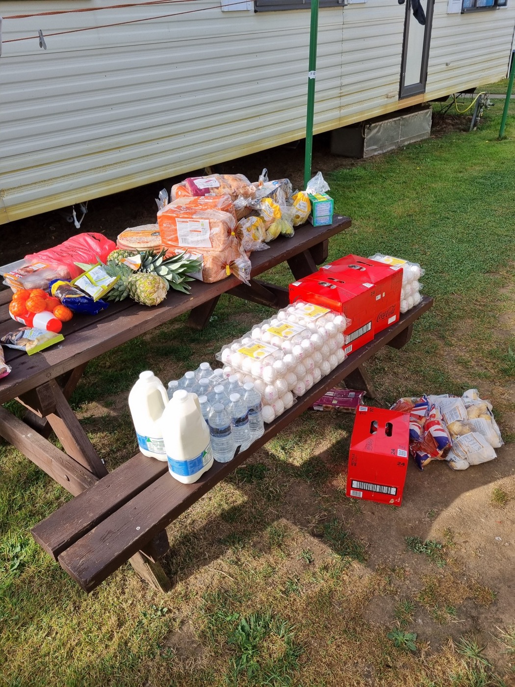 Деревянный стол для пикника у стены жилого каравана на Sheeplands Farm, на столе выложены продукты: хлеб, лотки с яйцами, бутылки молока, коробки